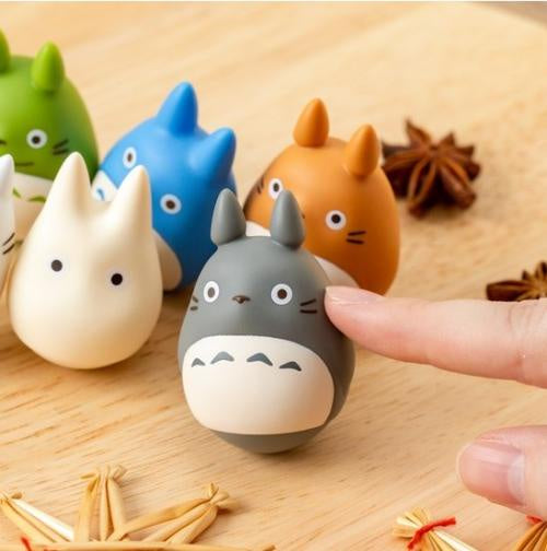 A close-up of a finger touching a small gray Totoro figurine on a light wood surface, with other colorful Totoro-style figures in the background and star anise props.